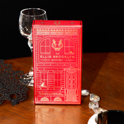 Red An Ellis Brooklyn Mystery Fragrance Discovery Set box on wooden table with diamond necklace, black and white bracelet, black lace mask, and stemmed glass, on black background.