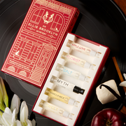 A red Ellis Brooklyn Mystery Fragrance Discovery Set box opened to show inside: Apple Love 1.5ml Vial, Florist 1.5ml Vial, Salt 1.5ml Vial, Myth 1.5ml Vial, Vanilla Milk 1.5ml Vial and Sweet 1.5ml Vial on a black vinyl record with vanilla beans, marshmallows, an apple, sandalwood and white flowers, on a wooden table.