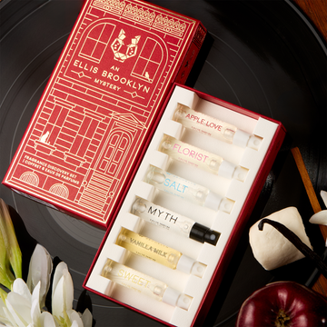 A red Ellis Brooklyn Mystery Fragrance Discovery Set box opened to show inside: Apple Love 1.5ml Vial, Florist 1.5ml Vial, Salt 1.5ml Vial, Myth 1.5ml Vial, Vanilla Milk 1.5ml Vial and Sweet 1.5ml Vial on a black vinyl record with vanilla beans, marshmallows, an apple, sandalwood and white flowers, on a wooden table.