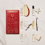 A red Ellis Brooklyn Mystery Fragrance Discovery Set box on white marble table with Apple Love 1.5ml Vial next to apple slice, Myth 1.5ml Vial next to white flower, Florist 1.5ml Vial next to white flower, Vanilla Milk 1.5ml Vial next to vanilla bean, Salt 1.5ml Vial next to sea salt, and Sweet 1.5ml Vial next to a marshmallow.