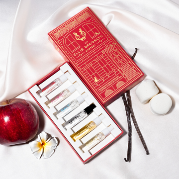 Coffret découverte de parfums Red An Ellis Brooklyn Mystery ouvert pour montrer l'intérieur avec les fioles de 1,5 ml Eau de Parfum Apple Love, Florist, Salt, Myth, Vanilla Milk et Sweet posées sur un drap en satin blanc avec des gousses de vanille, des guimauves, une pomme et une fleur blanche.