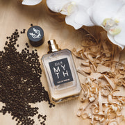 Myth 50ml Eau de Parfum avec le bouchon retiré sur une table en bois à côté de fleurs d'orchidée blanche, de graines d'ambarétte et de copeaux de bois de cèdre blanc