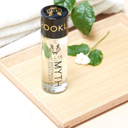 Huile corporelle parfumée Myth Spectacular 95 ml et fleur blanche sur une planche en bois, à côté de serviettes blanches pliées et de feuilles vertes, sur une table blanche.