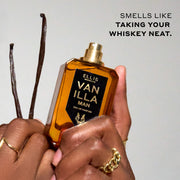 Model weaing gold rings holding Vanilla Man 50ml Eau De Parfum with cap removed in one hand, and vanilla beans in the other, on light grey background with black text that reads 