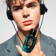 Modèle aux cheveux bruns portant une veste grise et des écouteurs, tenant le parfum Worth Avenue 50 ml Eau De Parfum contre le menton, sur fond blanc.