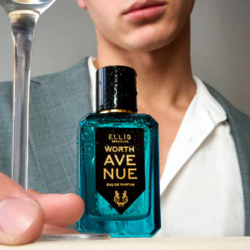 Model wearing grey jacket and white shirt holding stem of glass next to Worth Avenue 50ml Eau De Parfum covered in water droplettes on white table, on light grey background. | english_image