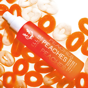Peaches 50ml Hair and Body Fragrance Mist without cap resting on white and orange candy peach rings, on white background.