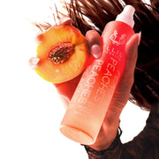 Model with brown hair and French manicure wearing black lace shirt holding half of a peach and Peaches 100ml Hair and Body Fragrance Mist without cap on white background.