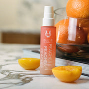 Peaches 50ml Hair and Body Fragrance Mist on white marble table next to peeled and cut peach halves, in front of a glass bowl full on oranges resting on a book, with a restaurant in the background.