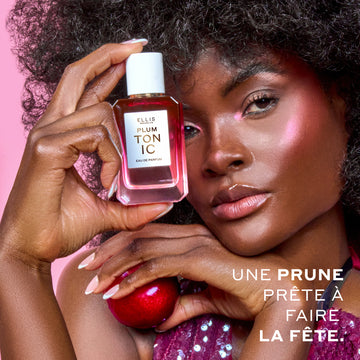 Une femme aux cheveux bruns portant un débardeur à sequins violet et du surligneur rose tient PLUM TONIC Eau de Parfum 50 ml contre sa tête d'une main et une prune de l'autre sur un fond rose avec un texte blanc qui dit « Une prune prête à faire la fête. »