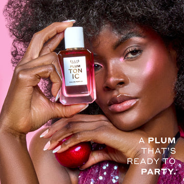 Woman with brown hair wearing a purple sequin tank top and pink highlighter holds PLUM TONIC Eau de Parfum 50ml against her head in one hand and a plum in the other hand on a pink background with white text that reads 