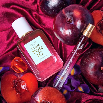 PLUM TONIC Eau de Parfum 50ml et PLUM TONIC Eau de Parfum 10ml à côté de prunes entières, de prunes tranchées, de pierres d'ambre et d'iris violets sur un tissu en velours rouge