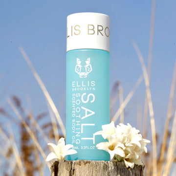 Huile corporelle parfumée apaisante au sel 95ml à côté de fleurs blanches sur un morceau de bois avec des dunes de plage et le ciel en arrière-plan.