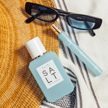 Salt 50ml Eau De Parfum and Salt 10ml Eau De Parfum Travel spray on tan straw hat and blue and white towel, next to black sunglasses.