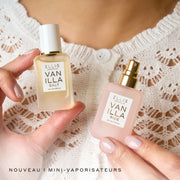 Des mini-flacons vaporisateurs de 7,5 mL de Vanilla Salt et Vanilla Rice Eau de Parfum tenus dans les mains d’une femme contre un haut en tricot blanc, avec du texte noir en bas à gauche qui indique « NOUVEAU ! MINI VAPORISATEURS ».