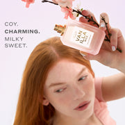 Woman with red hair wearing a pink shirt holds VANILLA RICE Eau de Parfum 50ml without cap and pink sakura blossoms above her head on a pink background with gray text that reads 