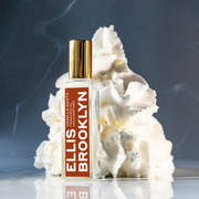 Vanilla Santo Fragrance Oil 15ml in front of swirl of whipped cream on glass table against grey background with smoke.