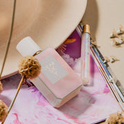 Le parfum Dear Sky 50ml Eau De Parfum et le spray voyage Dear Sky 10ml Eau De Parfum reposant sur une image de magazine violet et blanc et un chapeau beige avec des fleurs séchées, sur un fond beige.