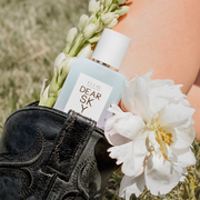 Model wearing black cowboy boot with Dear Sky 50ml Eau De Parfum and flowers tucked inside against calf, with green grass in background.