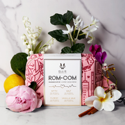 Red and pink pattered Rom-Com Fragrance Gift Set bag on white and grey marble backdrop with white, pink, purple, and yellow flowers, sandal wood, lemon, and bergamot.