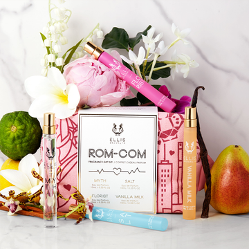 Florist 10ml Eau De Parfum Travel Spray on top of red and pink pattered Rom-Com Fragrance Gift Set bag, Myth 10ml Eau De Parfum Travel Spray, Salt 10ml Eau De Parfum Travel Spray, and Vanilla Milk 10ml Eau De Parfum Travel Spray on white and grey marble backdrop with white, pink, purple, and yellow flowers, sandal wood, lemon, pear, and bergamot.