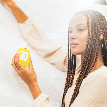 Model with brown hair and nude nail polish wearing an off-white long sleeve shirt, gold necklace, and gold rings, hold Sun Fruit 50ml Eau De Parfum in one hand against white background.