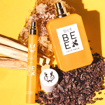 Bee 10ml Eau De Parfum travel Spray with cap removed and Bee 50ml Eau De Parfum with cap removed resting on piece of honeycomb, pieces on wood, and cocoa absolute, on yellow background.