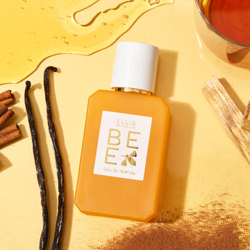 Bee 50ml Eau De Parfum on light yellow background with spilled honey, glass filled with honey, vanilla beans, cinnamon sticks, powdered cinnamon, and crystals.