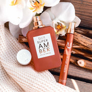 Super Amber 50ml Eau De Parfum with cap removed and Super Amber 10ml Eau De Parfum Travel Spray resting on pieces of wood and off-white knit blanket next to white flowers.