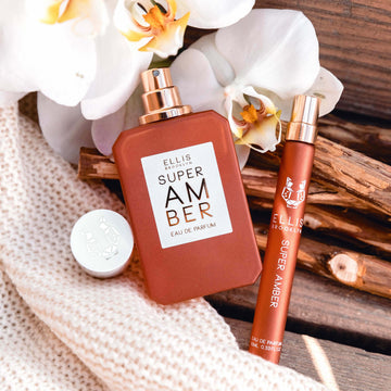 Super Amber 50ml Eau De Parfum with cap removed and Super Amber 10ml Eau De Parfum Travel Spray resting on pieces of wood and off-white knit blanket next to white flowers.