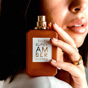 Model with brown hair, nude nail polish and nose ring wearing a white shirt and gold rings holding Super Amber 50ml Eau De Parfum with cap removed on shoulder.