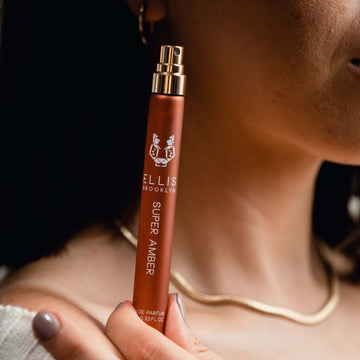 Model with brown hair and nude nail polish wearing white sweater, gold necklace, and gold earring holding Super Amber 10ml Eau De Parfum Travel Spray with cap removed on shoulder.