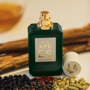 Apres 50ml Eau De Parfum with cap removed on white table with pieces of wood, berries, saffron, and orange drink in glass, on white background.