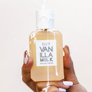 Model with white nail polish holding Vanilla Milk 50ml Eau De Parfum with milk being poured on bottle and hand on white background.