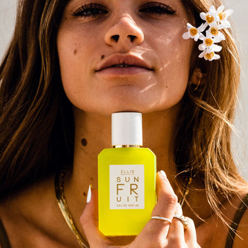 Model with brown hair and white nail polish wearing brown tank top, gold earrings, gold necklaces, gold rings, and white flowers in hair holds Sun Fruit 50ml Eau De Parfum below chin.