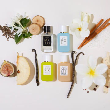 Myth 7.5ml Eau De Parfum Mini Dabber, Salt 7.5ml Eau De Parfum Mini Dabber, Sun Fruit 7.5ml Eau De Parfum Mini Dabber, and Vanilla Milk 7.5ml Eau De Parfum Mini Dabber surrounded by white flowers, cedar wood, ambrette seeds, sandal wood, sea salt, vanilla beans, cocoa shells, pear, and fig, on white background.