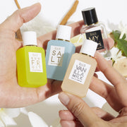 Model holding Myth 7.5ml Eau De Parfum Mini Dabber, Salt 7.5ml Eau De Parfum Mini Dabber, Sun Fruit 7.5ml Eau De Parfum Mini Dabber, and Vanilla MIlk 7.5ml Eau De Parfum Mini Dabber in both hands over white flowers, sandalwood, green leaves, and cedarwood, on white background.