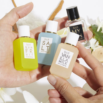 Model holding Myth 7.5ml Eau De Parfum Mini Dabber, Salt 7.5ml Eau De Parfum Mini Dabber, Sun Fruit 7.5ml Eau De Parfum Mini Dabber, and Vanilla MIlk 7.5ml Eau De Parfum Mini Dabber in both hands over white flowers, sandalwood, green leaves, and cedarwood, on white background.