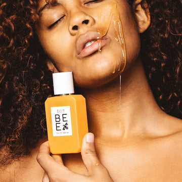 Model with brown curly hair holding Bee 50ml Eau De Parfum against jaw with honey dripping down cheek, jaw and lips, onto hand.