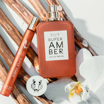 Super Amber 50ml Eau De Parfum with cap removed and Super Amber 10ml Eau De Parfum Travel Spray resting on pieces of wood next to white flowers, on white background.