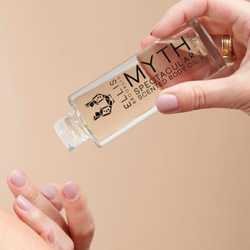 Model with nude nail polish wearing gold rings holding Model holding Myth 95ml Spectacular Scented Body Oil with out cap in one hand while dispensing product onto other hand.