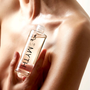 Model holding Myth Spectacular Scented Body Oil 95ml with cap removed near chest on white background.