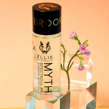 Myth Spectacular Scented Body Oil 95ml on glass block in front of pink flowers, glass block, and glass cylinder, on orange background.