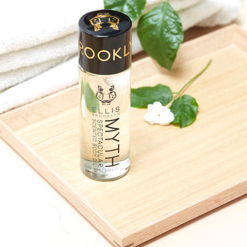 Myth Spectacular Scented Body Oil 95ml and white flower on wooden board, next to folded white towels and green leaves, on white table.