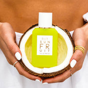 Model with white nail polish, wearing assorted gold and silver rings, white pants, and a white long sleeved shirt holding Sun Fruit 50ml Eau De Parfum in a coconut with both hands against bare stomach.