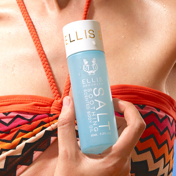 Model wearing orange, pink, and brown bikini top holding Salt 95ml Soothing Scented Body Oil one hand near chest, on blue background.
