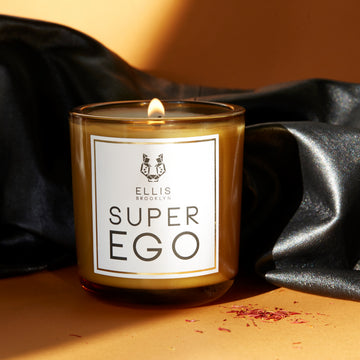 Superego 6.5 oz. Terrific Scented Candle with wick lit on orange backdrop with saffron and black leather.