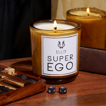 Superego 6.5 oz. Terrific Scented Candle on wooden table next to game with dice and dice on table, in front of brown book, mirror, and black fabric.