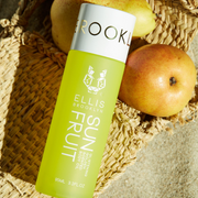 Sun Fruit 95ml Glistening Golden Scented Body Oil resting on pears and wicker bag on sand.