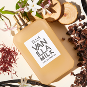 Vanilla Milk 50ml Eau De Parfum without cap on off white background surrounded by cedar wood, white flowers, vanilla beans, saffron, and cocoa shells.